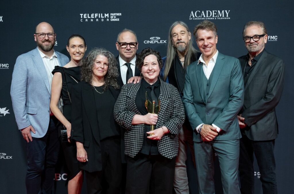 A group of eight people (three women and five men) stand at the Canadian Screen Awards winners' wall. The woman in the centre is holding a trophy. Everyone is wearing suits or dresses, and they are smiling.