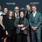 A group of eight people (three women and five men) stand at the Canadian Screen Awards winners' wall. The woman in the centre is holding a trophy. Everyone is wearing suits or dresses, and they are smiling.