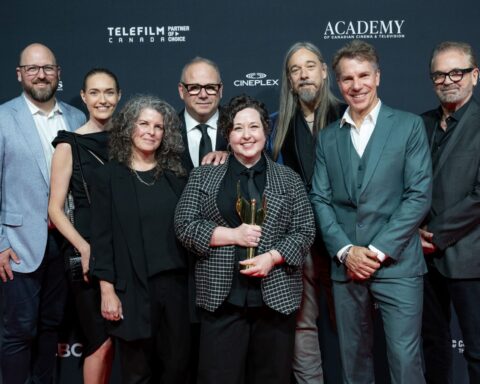 A group of eight people (three women and five men) stand at the Canadian Screen Awards winners' wall. The woman in the centre is holding a trophy. Everyone is wearing suits or dresses, and they are smiling.