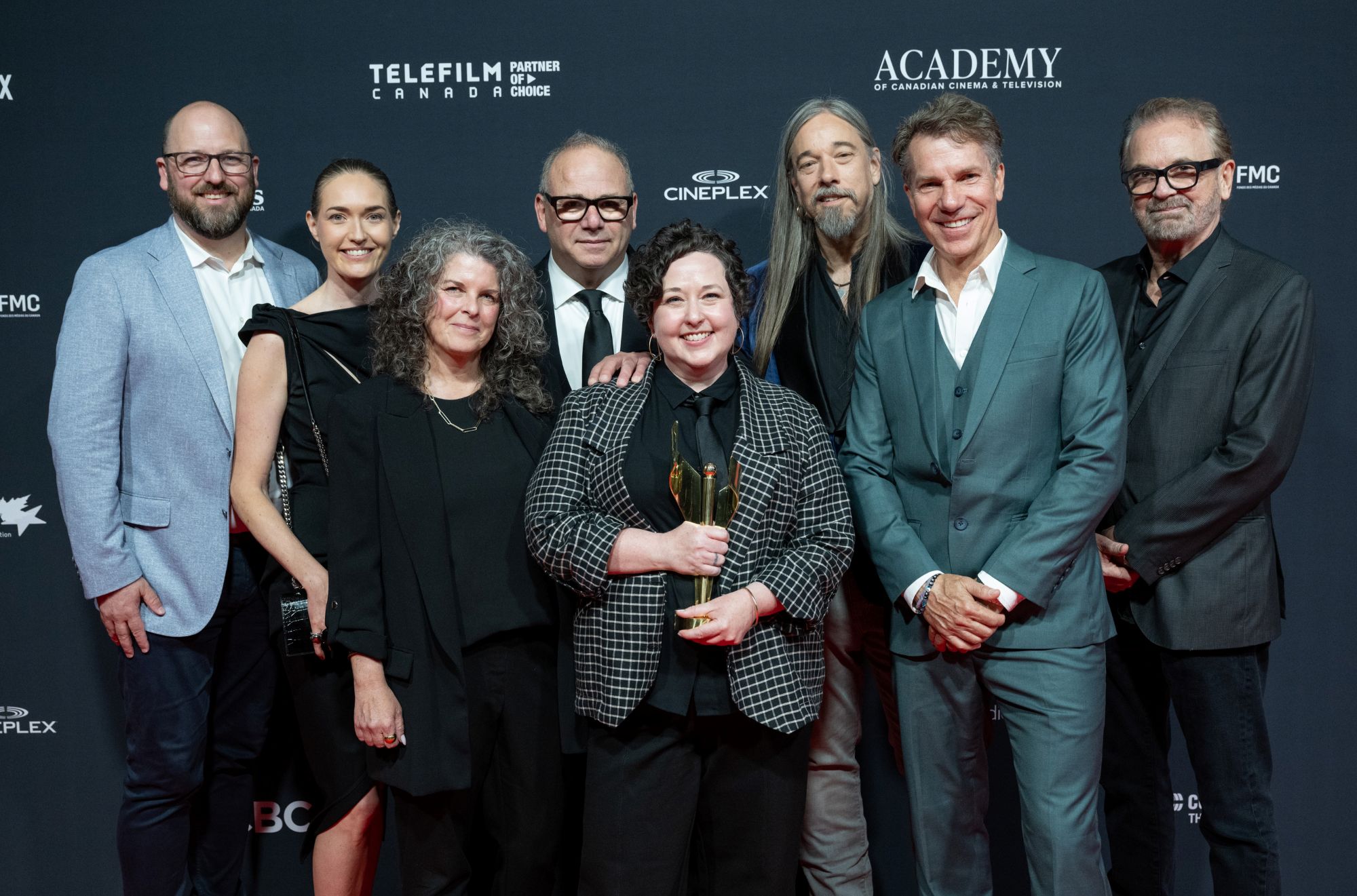 A group of eight people (three women and five men) stand at the Canadian Screen Awards winners' wall. The woman in the centre is holding a trophy. Everyone is wearing suits or dresses, and they are smiling.