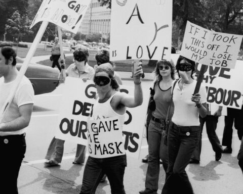 A black and white photo of a protest march. Several people are wearing masks. They are holding placards with messages including "If I took this off, I would lose my job."