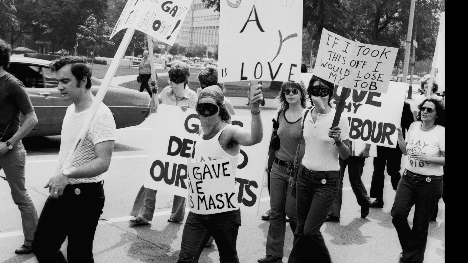 A black and white photo of a protest march. Several people are wearing masks. They are holding placards with messages including "If I took this off, I would lose my job."