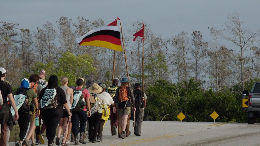 A group of people carrying a Miccosukee flag and wearing banners that say “protect the land” and “defend sacred land and water” march down a road in the Everglades