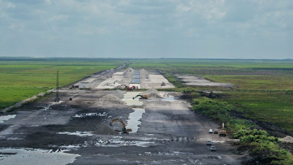 Excavators dig new canals and infrastructure in the Everglades. The green landscape is ravaged by mud and craters. The sky is blue.