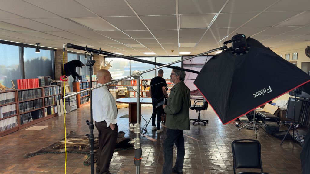 Two men stand talking in front of a film interview set up. There is equipment in the background.
