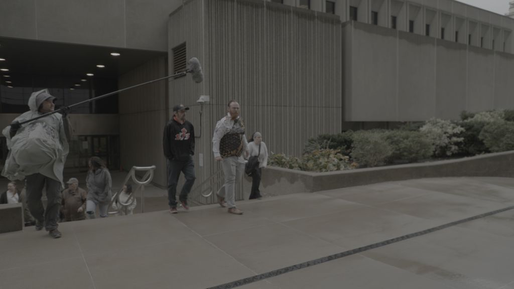 A crowd of people leaves a courthouse. They are visibly upset and there is a TV crewwith a boom mic walking beside them.