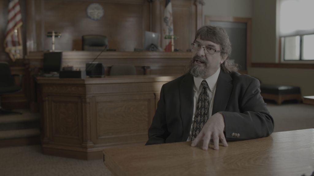 A man in a suit sits in a courtroom giving an interview. His hand is splayed on a table as he speaks passionately.