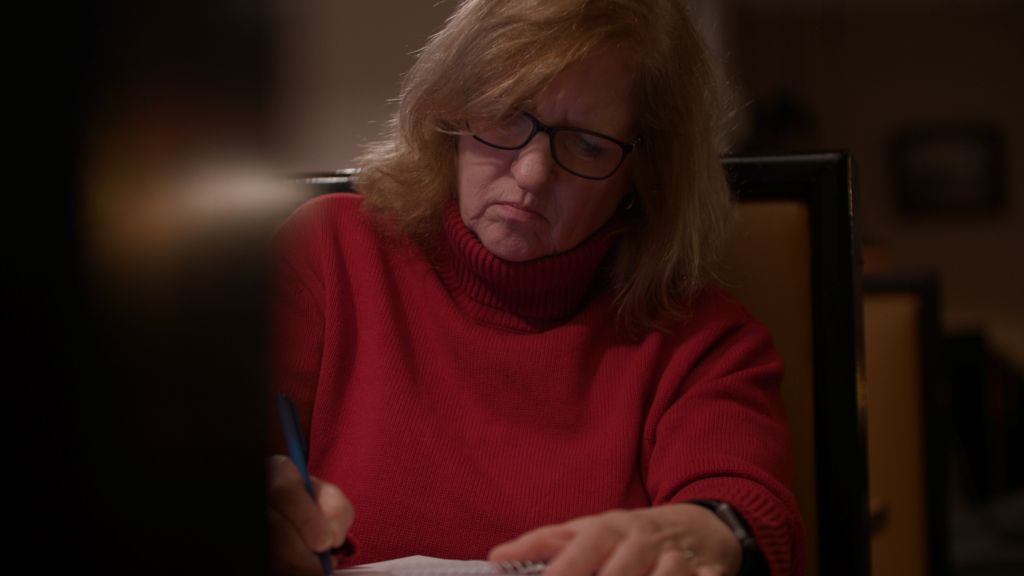 A woman with blonde hair and dark plastic glasses sits in a diner booth, writing with a pen and paper. She is wearing a red sweater.