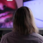 The view from behind of a man with long hair watching a video screen. The screen includes a partial face of a man with a moustache and beard.