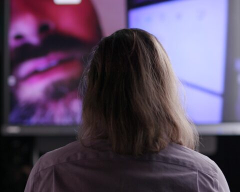 The view from behind of a man with long hair watching a video screen. The screen includes a partial face of a man with a moustache and beard.