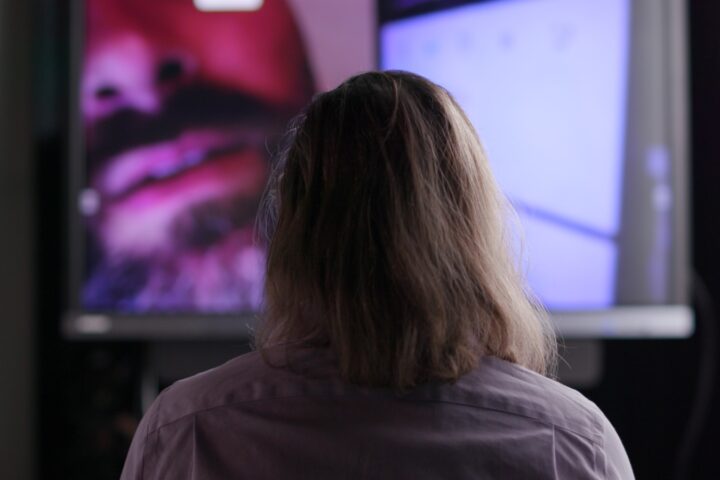 The view from behind of a man with long hair watching a video screen. The screen includes a partial face of a man with a moustache and beard.