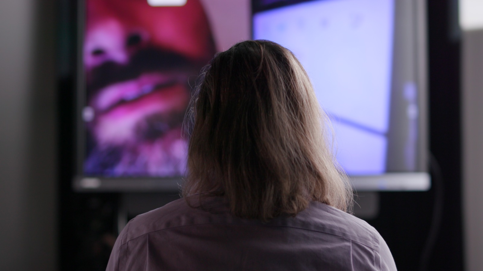 The view from behind of a man with long hair watching a video screen. The screen includes a partial face of a man with a moustache and beard.