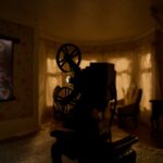 An old family home movie is projected onto the living room of a doll house. The room is dark and the projector is in the foreground. There are widows covered with drapes in the background.