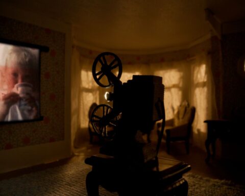 An old family home movie is projected onto the living room of a doll house. The room is dark and the projector is in the foreground. There are widows covered with drapes in the background.