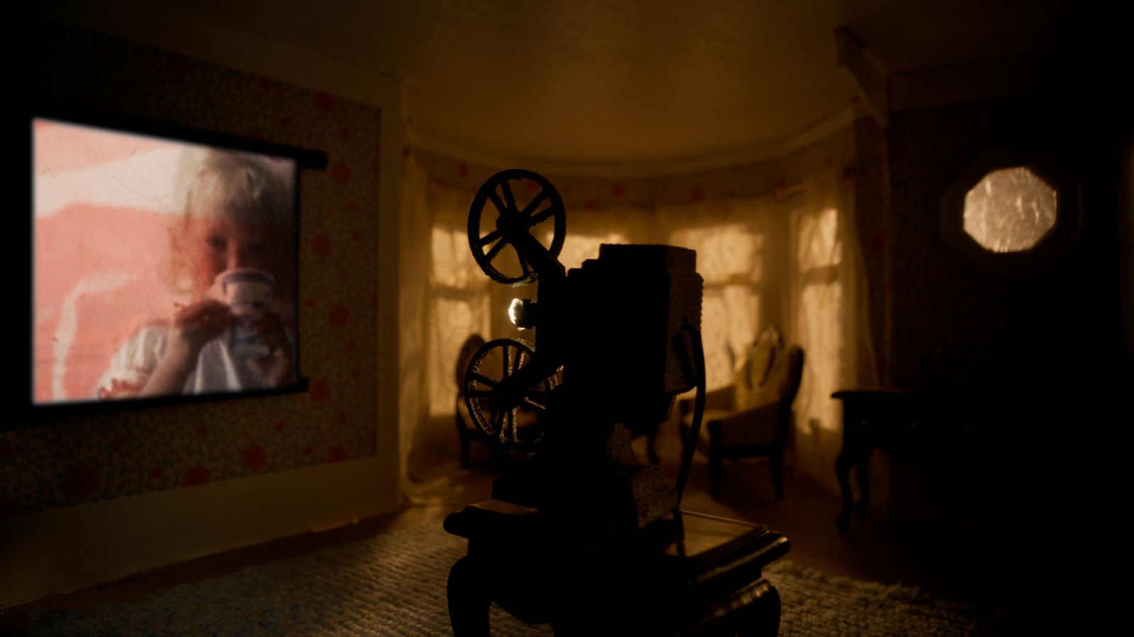 An old family home movie is projected onto the living room of a doll house. The room is dark and the projector is in the foreground. There are widows covered with drapes in the background.