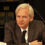 Wikileaks founder Julian Assange sits at a desk. He has white hair and is wearing a brown suit with a white shirt and black tie.