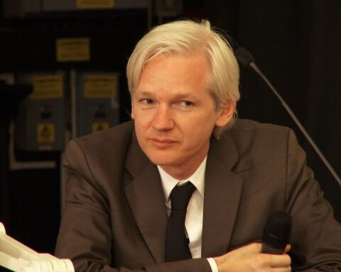 Wikileaks founder Julian Assange sits at a desk. He has white hair and is wearing a brown suit with a white shirt and black tie.