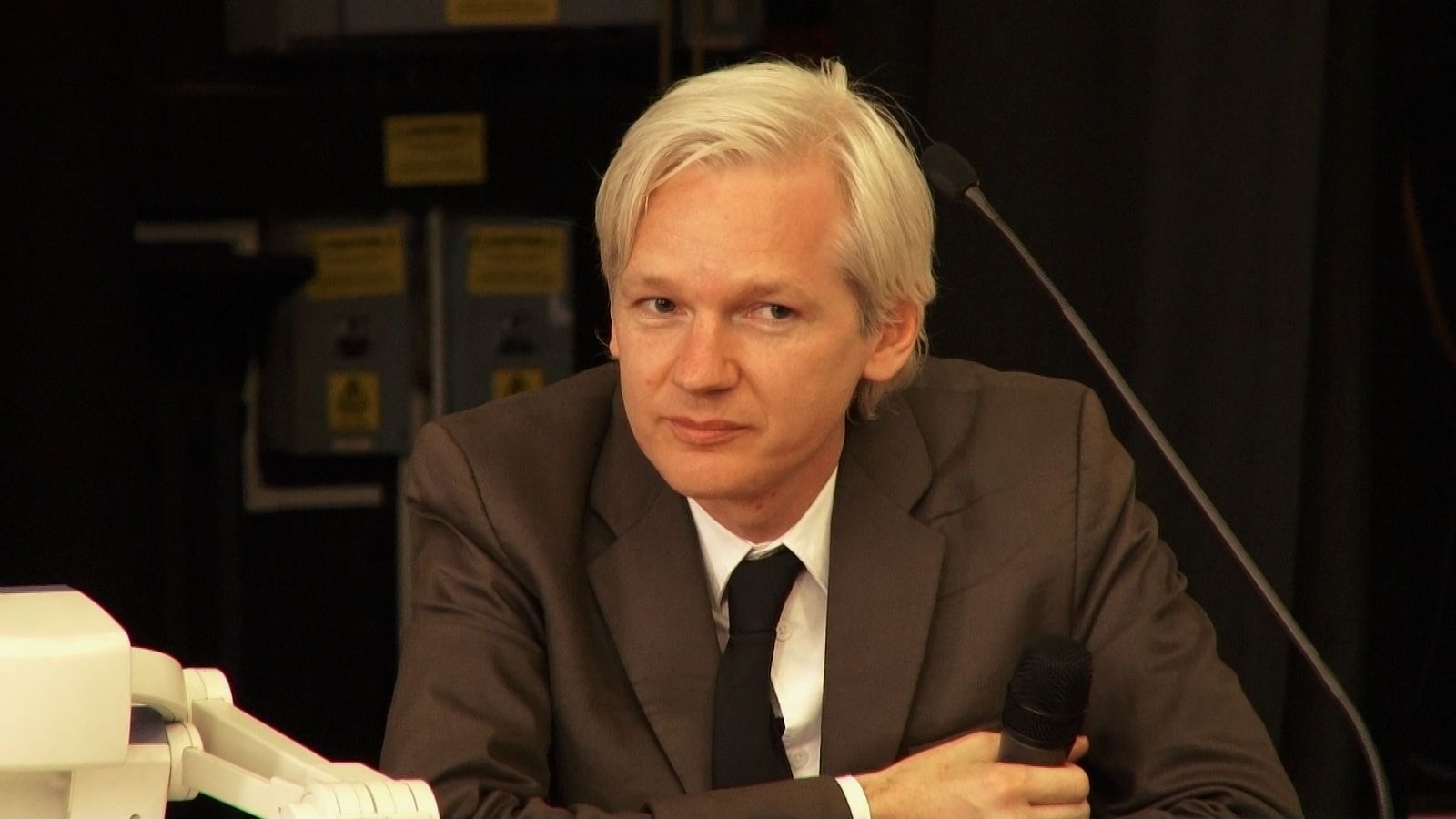 Wikileaks founder Julian Assange sits at a desk. He has white hair and is wearing a brown suit with a white shirt and black tie.
