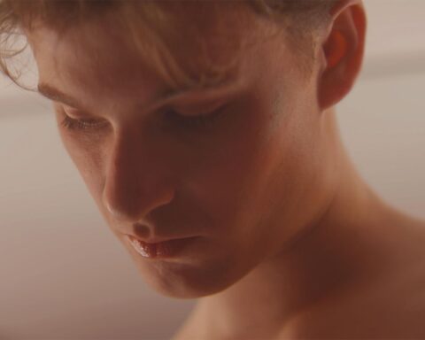 A Caucasian teenage boy with red hair is viewed in close-up. He is looking down and is wearing a black and white tank top and light pink lip gloss.