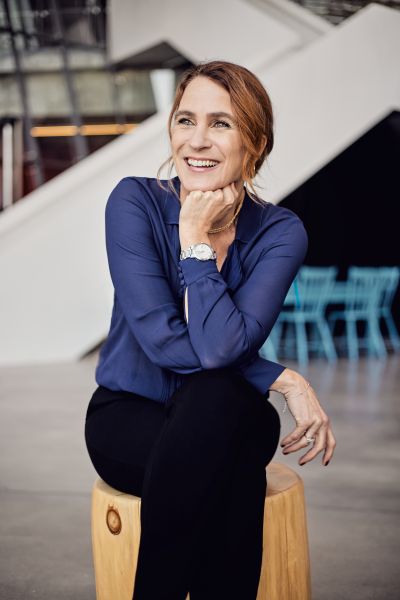 Suzanne Guèvremont is a white woman with blond hair. She is sitting cross-legged with a hand supporting her chin. She is wearing a purple blouse and black pants.