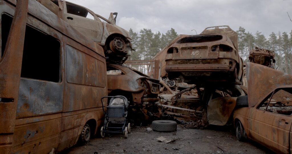 An abandoned stroller sits amid a heap of rusted cars destroyed by Russian violence in Ukraine.