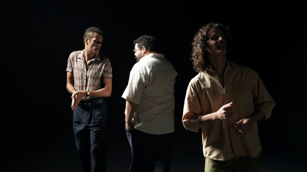 Three actors appear on a black stage. They have spotlights on them and are interacting.