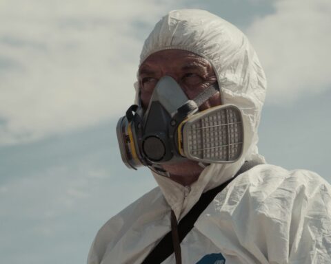 A person is seen from the shoulder up wearing a nuclear radiation protective gear and a breathing mask. There is a blue sky with clouds behind them.