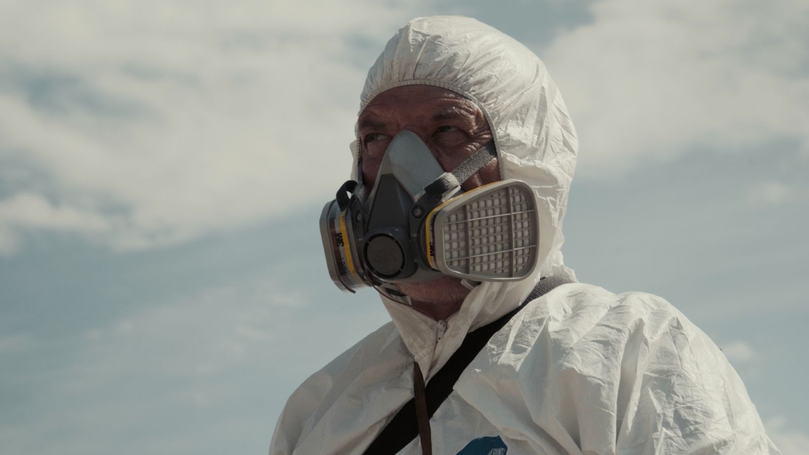 A person is seen from the shoulder up wearing a nuclear radiation protective gear and a breathing mask. There is a blue sky with clouds behind them.