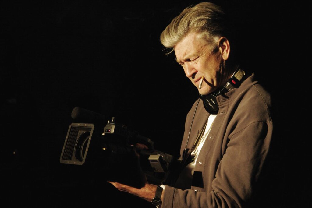 Filmmaker David Lynch holds a camera at waist-level and looks down into the viewfinder. He is wearing a brown hoodie with a white t-shirt, and a cigarette is dangling from his mouth. He has grey hair and is photographed against a black background.