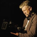 Filmmaker David Lynch holds a camera at waist-level and looks down into the viewfinder. He is wearing a brown hoodie with a white t-shirt, and a cigarette is dangling from his mouth. He has grey hair and is photographed against a black background.