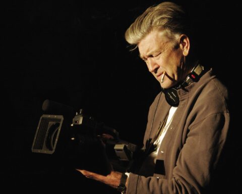 Filmmaker David Lynch holds a camera at waist-level and looks down into the viewfinder. He is wearing a brown hoodie with a white t-shirt, and a cigarette is dangling from his mouth. He has grey hair and is photographed against a black background.