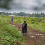 A woman stands in a green field with a dirt path. Her back is facing the camera. The sky is grey and cloudy.