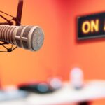 A podcast microphone is suspended by a boom. There is a black On Air sign with orange lettering. The walls of the studio are bright orange and there is a table with a laptop in the background.