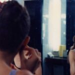 A ballet dancer is seated at a mirror. She is smiling and putting on an earring.