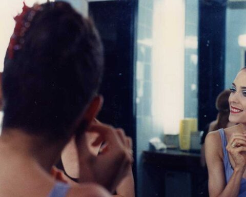A ballet dancer is seated at a mirror. She is smiling and putting on an earring.