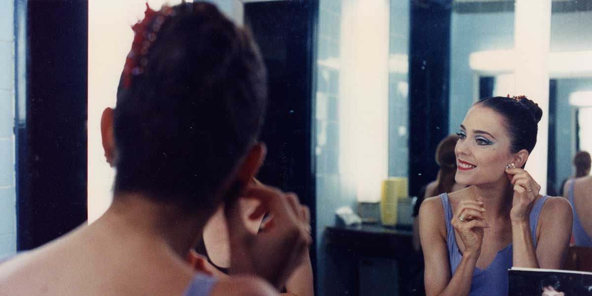 A ballet dancer is seated at a mirror. She is smiling and putting on an earring.