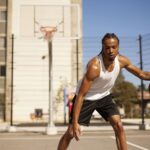 A young Black male dribbles a basketball on an outdoor court. He is wearing a white tank top, and black shorts with his hair in braids. There is a basketball hoop behind him, and the court is surrounded by apartment buildings and a blue sky.