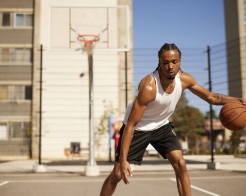 A young Black male dribbles a basketball on an outdoor court. He is wearing a white tank top, and black shorts with his hair in braids. There is a basketball hoop behind him, and the court is surrounded by apartment buildings and a blue sky.