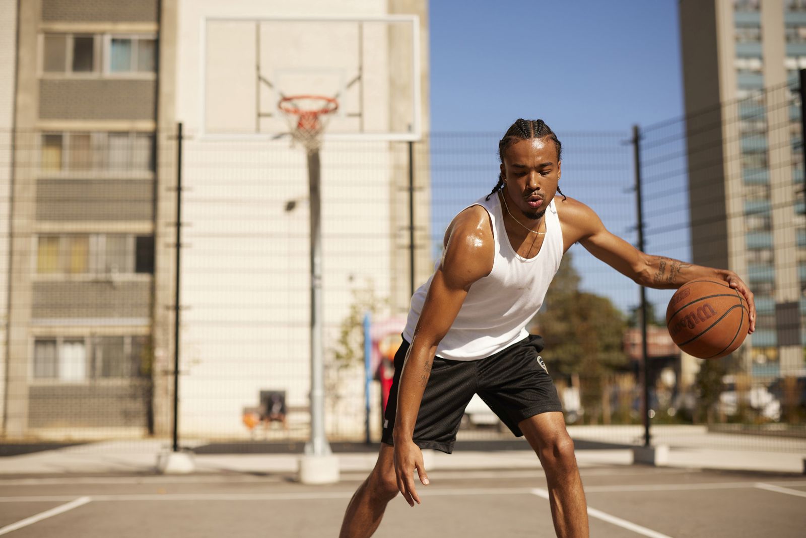 A young Black male dribbles a basketball on an outdoor court. He is wearing a white tank top, and black shorts with his hair in braids. There is a basketball hoop behind him, and the court is surrounded by apartment buildings and a blue sky.