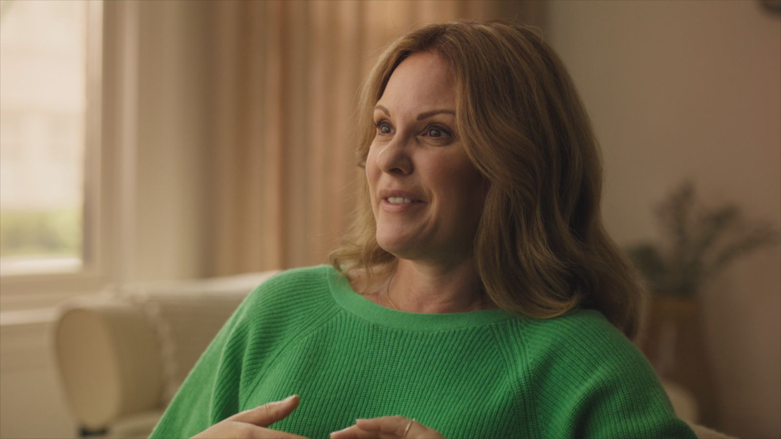Heather is a white woman with blond hair and brown eyes. She is seated giving an interview. She is wearing a green sweater and seated in front of a window with white curtains.