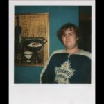 A vintage polaroid photo of actor John Candy. He is wearing a Toronto Maple Leafs jersey and smiling. Behind him is a blue wall with a yellow painting.