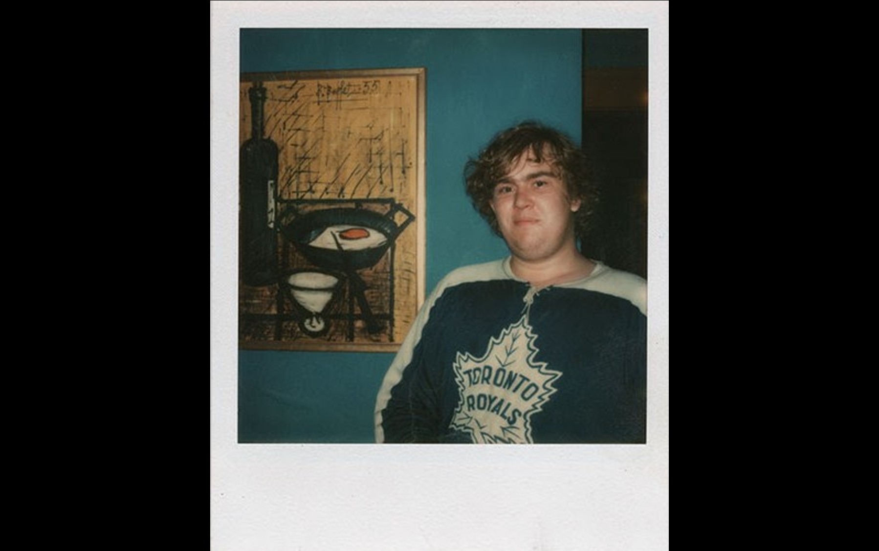 A vintage polaroid photo of actor John Candy. He is wearing a Toronto Maple Leafs jersey and smiling. Behind him is a blue wall with a yellow painting.