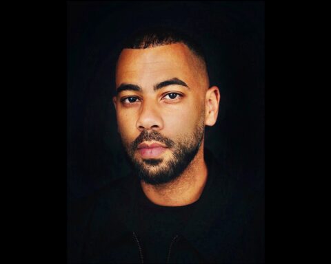 Julian Carrington is a Black man with a beard and moustache, and short cropped hair. He is wearing a black shirt and photographed in portrait.