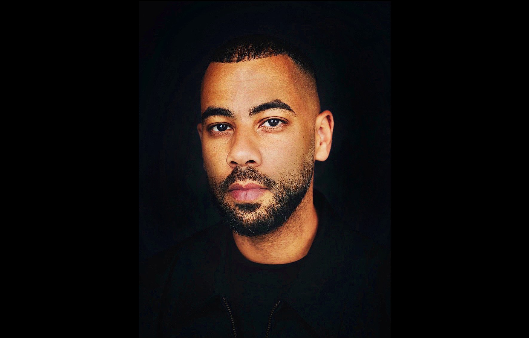 Julian Carrington is a Black man with a beard and moustache, and short cropped hair. He is wearing a black shirt and photographed in portrait.