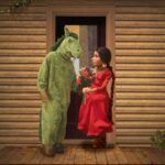 A man dressed in a costume as a green horse stands on his back hooves and talks to a puppet woman in a red dress. She is holding a bouquet of multicoloured flowers. They are standing on the deck of a log cabin.