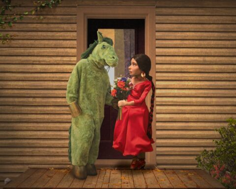 A man dressed in a costume as a green horse stands on his back hooves and talks to a puppet woman in a red dress. She is holding a bouquet of multicoloured flowers. They are standing on the deck of a log cabin.