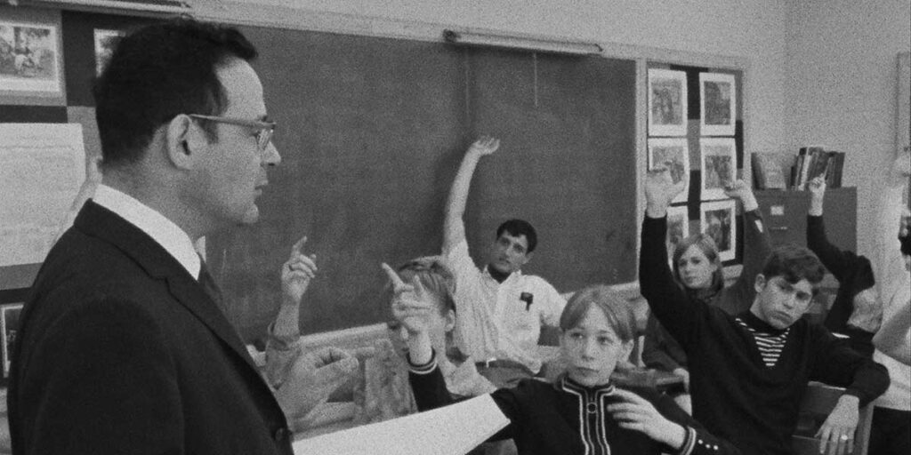 A black and white photo of a high school class in the 1960s. A teacher is in the foreground of the frame. He is looking at his class of students, who face him with their hands raised in the air.