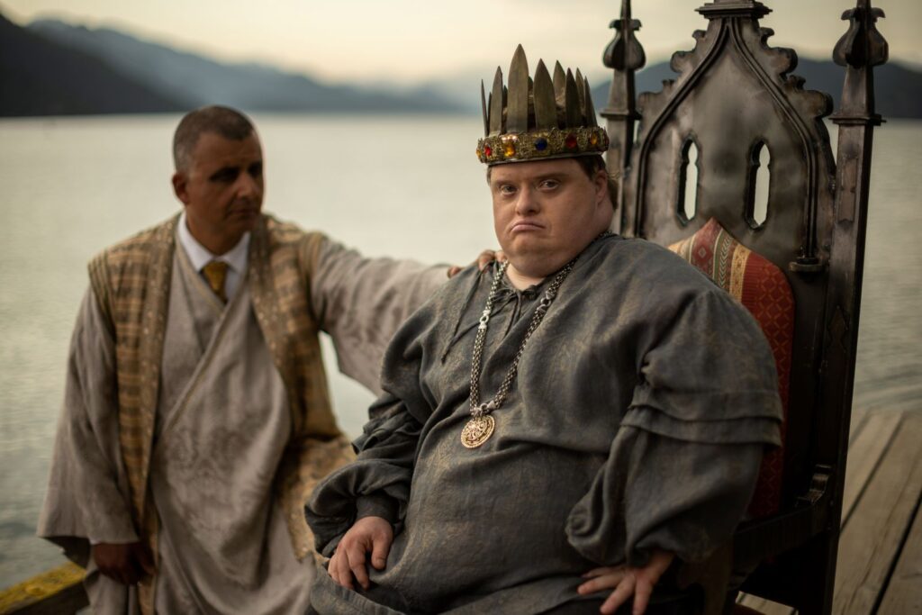 Niall McNeil, an actor with Down syndrome, sits on a throne wearing a crown and medieval costume. An actor stands behind him, wearing a suit and robe, with his hand leaning on the throne.