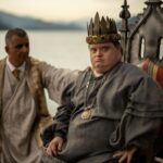 Niall McNeil, an actor with Down syndrome, sits on a throne wearing a crown and medieval costume. An actor stands behind him, wearing a suit and robe, with his hand leaning on the throne.