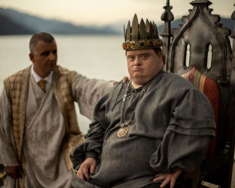 Niall McNeil, an actor with Down syndrome, sits on a throne wearing a crown and medieval costume. An actor stands behind him, wearing a suit and robe, with his hand leaning on the throne.